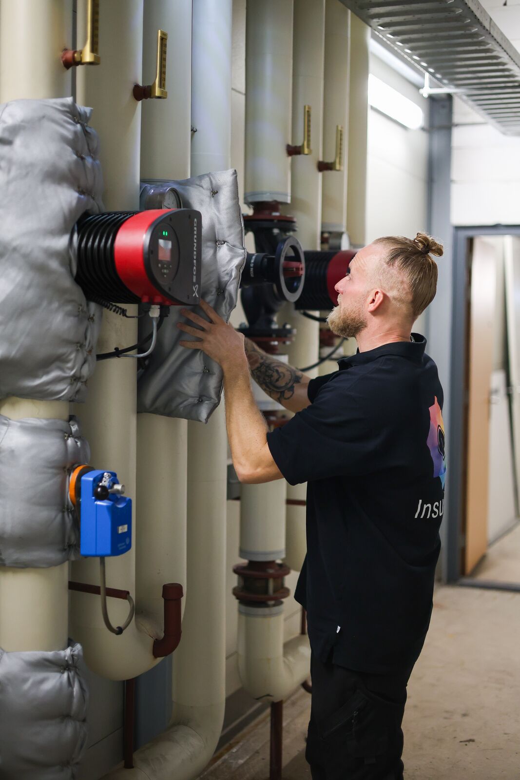 InsulSafe-monteur past een isolatiematras aan op een circulatiepomp in de technische ruimte